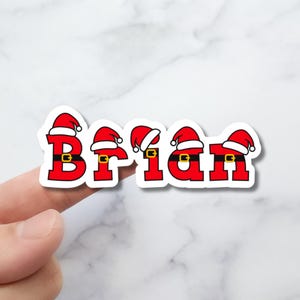 May include: A red and white Christmas-themed sticker with the name "Brian" spelled out in large letters. Each letter has a Santa hat on top and a black belt with a gold buckle.