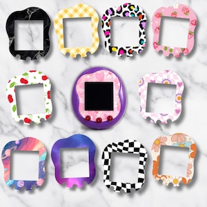 May include: A collection of decorative frames with various patterns, including marble, gingham, leopard print, strawberries, cherries, flowers, and a checkerboard design. A purple handheld digital pet game is in the center. The frames are designed to fit the game.