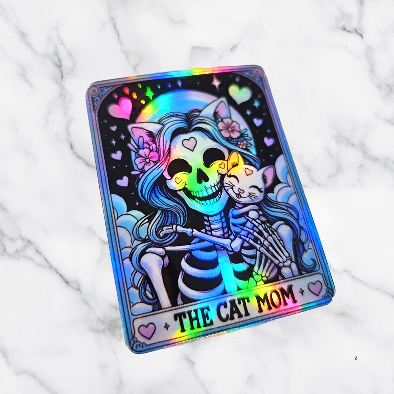 May include: Holographic sticker featuring a skeleton with blue hair and floral accents, embracing a cat. The design includes hearts, a rainbow, and the text "THE CAT MOM" in a decorative frame. The sticker has a shiny, iridescent finish.