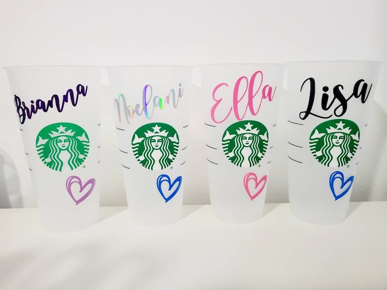 K&ouml;nnte beinhalten: Vier wei&szlig;e wiederverwendbare Becher mit schwarzem, lila, rosa und gr&uuml;nem Text. Die Becher haben ein gr&uuml;nes Starbucks-Logo mit dem Gesicht einer Frau und einem Herz in einer anderen Farbe auf jedem Becher. Die Namen Brianna, Melani, Ella und Lisa sind auf den Bechern geschrieben.