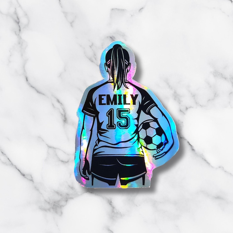 May include: Holographic sticker of a soccer player, viewed from the back, holding a soccer ball. The player wears a jersey with the name "EMILY" and the number "15". The sticker has a shiny, iridescent finish, with a marble background.
