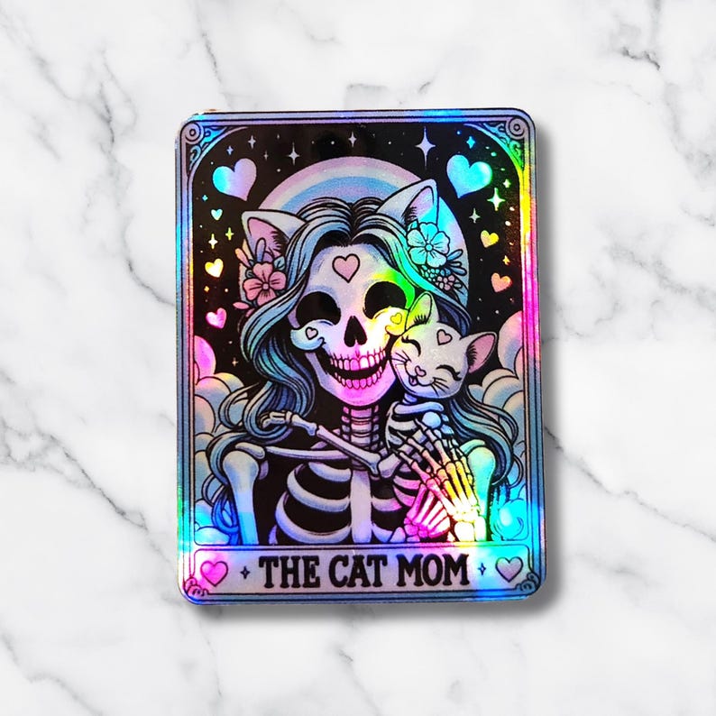 May include: A holographic sticker featuring a skeleton with long blue hair, cat ears, and flowers, embracing a skeleton cat. The design includes a rainbow, hearts, and the text "THE CAT MOM".