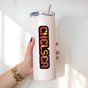 May include: A light pink insulated tumbler with a silver straw and a lid. The word "CHELSEA" is printed vertically in a pixelated, brown font with colorful accents. The tumbler is held by a person with a gold bracelet.