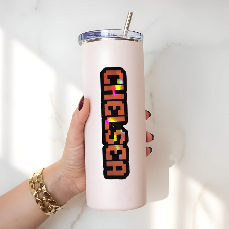 May include: A light pink insulated tumbler with a silver straw and a lid. The word "CHELSEA" is printed vertically in a pixelated, brown font with colorful accents. The tumbler is held by a person with a gold bracelet.