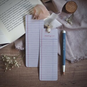 Könnte beinhalten: Zwei weiße Lesezeichenkarten mit blauen Linien und roten Rändern. Die obere Karte trägt den Text "Reading List." in roter Tinte. Die Karten liegen auf einer Holzoberfläche mit einem weißen Stift und einem Strauß weißer Blumen.