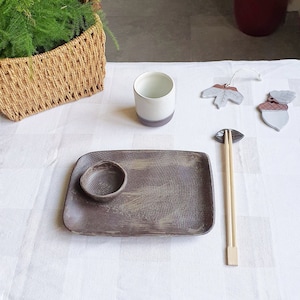 Puede incluir: Un plato cuadrado de cerámica marrón con un cuenco más pequeño en la parte superior, una taza de cerámica blanca, un par de palillos de madera y dos adornos de cerámica en forma de bellota y hoja sobre un mantel blanco.