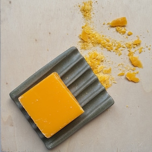 Può includere: Una saponetta di colore arancione brillante si trova su un portasapone in ceramica grigia. Il portasapone ha un design rigato ed è di forma rettangolare. La saponetta è leggermente rialzata rispetto al portasapone.