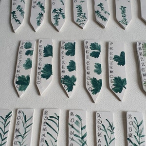 Può includere: Un set di 10 segnalini per piante in ceramica bianca con foglie dipinte di verde e lettere nere. I segnalini sono etichettati con i nomi delle erbe: Timo, Prezzemolo, Rosmarino.