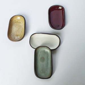 May include: Four small oval ceramic dishes in different colours. The dishes are arranged in a row, with the red dish at the top, the white dish in the middle, and the green and yellow dishes at the bottom.