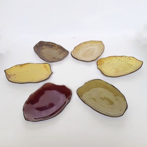Könnte beinhalten: Sechs Keramikteller in verschiedenen Farben und Formen. Die Teller sind alle handgefertigt und haben ein rustikales, erdiges Aussehen. Sie sind perfekt zum Servieren von Vorspeisen, Desserts oder Snacks.