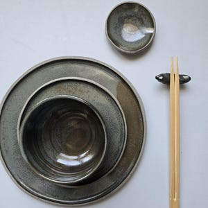 Könnte beinhalten: Ein Keramik-Geschirrset in Grau- und Brauntönen, bestehend aus Tellern, Schalen und einer kleinen Schale. Das Set wird durch ein Paar Bambus-Essstäbchen und eine Essstäbchenablage ergänzt. Das Geschirr hat eine gesprenkelte, strukturierte Oberfläche.