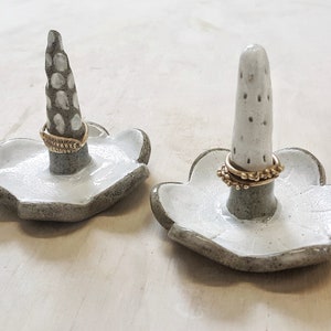 Può includere: Due porta anelli in ceramica bianca con accenti grigi. Ogni porta anelli ha un cono rialzato e testurizzato al centro e una base a forma di fiore. Un porta anelli ha un anello dorato sul cono, mentre l'altro ha due anelli dorati.