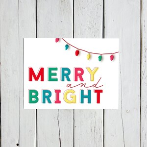 Könnte beinhalten: Eine weiße rechteckige Karte mit den Worten "MERRY and BRIGHT" in bunten Blockbuchstaben. Oben befindet sich eine Lichterkette mit roten, türkisfarbenen, gelben und rosa Lichtern. Die Karte liegt auf einer weißen Holzoberfläche.