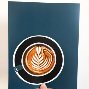 May include: A print featuring a latte art design in a black coffee cup with a teal handle, set against a dark teal background. The coffee art is a detailed leaf pattern in shades of brown and cream. The cup sits on a white circle.