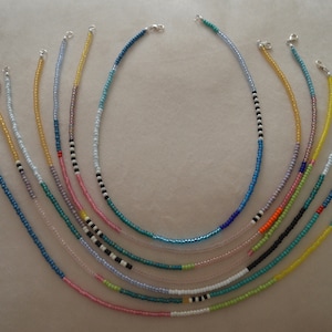 May include: A collection of 10 colorful beaded necklaces with silver clasps. The necklaces are arranged in a fan shape, with the longest necklace at the top and the shortest necklace at the bottom. The necklaces are made with small, round beads in various colors, including blue, green, yellow, pink, and white.