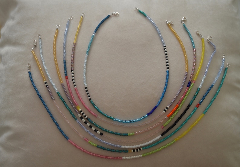 May include: A collection of 10 colorful beaded necklaces with silver clasps. The necklaces are arranged in a fan shape, with the longest necklace at the top and the shortest necklace at the bottom. The necklaces are made with small, round beads in various colors, including blue, green, yellow, pink, and white.