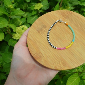 May include: A colorful beaded bracelet with black and white stripes, turquoise, yellow, and pink beads. The bracelet is on a wooden surface.
