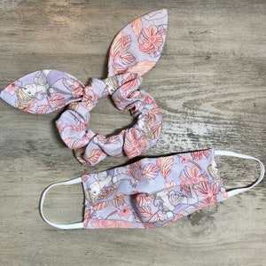 May include: A purple and pink floral print scrunchie and face mask set. The scrunchie is made of a soft, stretchy fabric and has a bow at the top. The face mask is made of a similar fabric and has elastic ear loops.