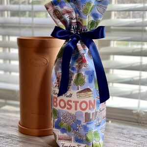 May include: A white fabric wine bottle bag with a blue ribbon tied around it. The bag is printed with a watercolor illustration of Boston landmarks, including the Freedom Trail, Faneuil Hall, and the Boston skyline. The text "BOSTON" is printed in red letters.