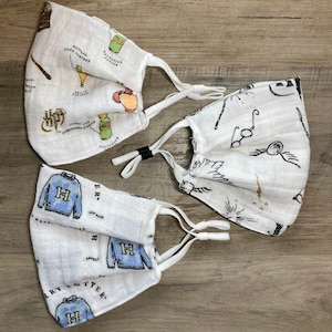 May include: Three white cotton face masks with Harry Potter themed designs. The first mask has a design of potions and magical objects. The second mask has a design of glasses, a wand, and a phoenix. The third mask has a design of a blue sweater with the letter H on it and the words "Harry Potter".