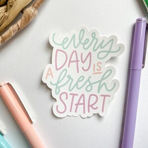 Peut inclure: Un autocollant avec le texte "Every day is a fresh start" en lettres roses, vertes et bleues sur un fond blanc.