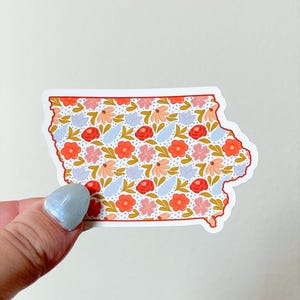 Op de afbeelding: Een sticker in de vorm van de staat Iowa, gevuld met een bloemenpatroon. De bloemen zijn rood, roze en perzik, met blauwe bladeren en gouden accenten. De sticker heeft een witte rand en wordt door een hand vastgehouden.