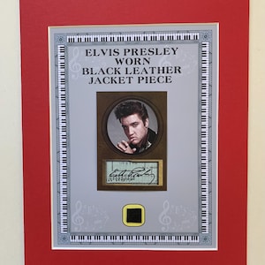 Puede incluir: Exhibición enmarcada con Elvis Presley. La imagen incluye un retrato de Elvis, un trozo de su chaqueta de cuero negro y su firma. La exhibición está sobre un fondo gris con notas musicales y teclas de piano, enmarcada en rojo.