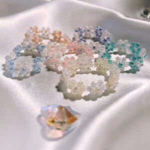 Puede incluir: Un conjunto de seis anillos de cuentas con diseños en forma de flor en varios colores pastel, incluyendo azul claro, rosa y amarillo. Los anillos están hechos de pequeñas cuentas redondas y están dispuestos en un círculo.