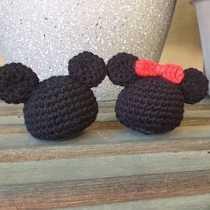 Puede incluir: Dos personajes de Mickey Mouse de crochet negro, uno con un lazo rojo, sentados sobre una superficie de madera.