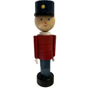 May include: A wooden toy soldier figurine. The figurine has a navy blue hat with a yellow dot, a light wood head, a red jacket, blue pants, and a black base. The toy soldier has a smiling face and white hands.