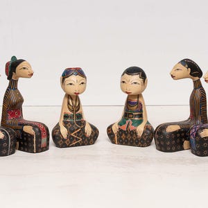 以下が含まれることがあります： 座っている姿の6体の手彫りの木製フィギュア。フィギュアは首が長く、カラフルな模様の服を着ています。カラーパレットには、緑、赤、黒、金が含まれます。フィギュアは白い背景に並んでいます。