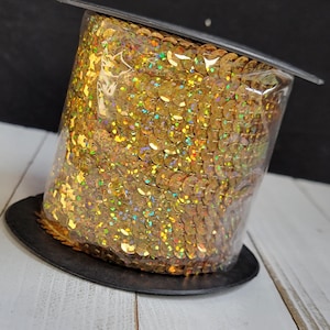 Puede incluir: Un carrete de lentejuelas doradas, bien empaquetadas y visibles a través de plástico transparente. Las lentejuelas son redondas y reflejan la luz con un efecto brillante e iridiscente. El carrete se asienta sobre una base negra, sobre un fondo negro y una superficie de madera blanca.