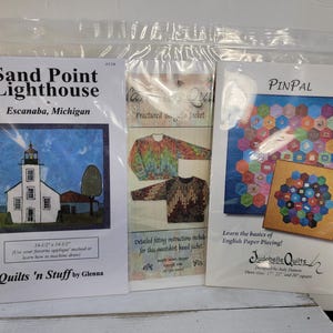 Puede incluir: Tres patrones de acolchado empaquetados. El patrón "Sand Point Lighthouse" presenta una ilustración de un faro. El patrón "Fractured Jacket" muestra diseños de chaquetas. El patrón "PINPAL" muestra un diseño de edredón hexagonal colorido. Todos los patrones están en plástico transparente.