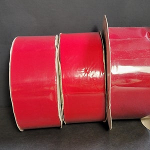 May include: Three rolls of red ribbon are shown against a dark backdrop. The ribbon is a solid, vibrant red colour and appears glossy. The rolls are different sizes, with the largest roll on the right. The ribbon is likely for crafts.