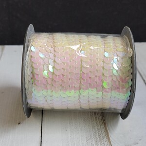 Könnte beinhalten: Eine Rolle schillernder Pailletten in einem blassrosa, gelben und grünen Farbschema. Die runden Pailletten sind dicht gepackt und erzeugen einen schimmernden Effekt. Die Rolle besteht aus grauem Material und liegt auf einer weißen Holzoberfläche.