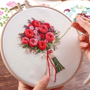 May include: An embroidery hoop with a floral design. The design features a bouquet of red roses, pink buds, and green foliage. The bouquet is tied with a bundle of green and beige threads. The embroidery is set against a white fabric.