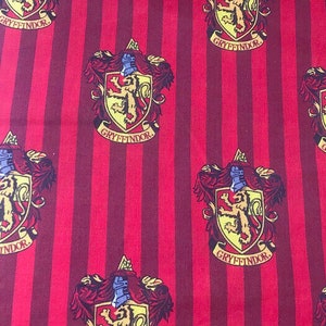 Op de afbeelding: Rood en bordeaux gestreepte stof met een herhalend patroon van het Gryffindor-wapen. Het wapen toont een leeuw, een helm en het woord "Gryffindor" op een spandoek. Geschikt voor diverse knutselprojecten.