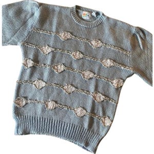 May include: A light blue knit sweater with a crew neck and long sleeves. The sweater features horizontal rows of diamond-shaped, off-white embellishments. The sleeves have a slight puff at the shoulders. The sweater has a ribbed hem and cuffs.