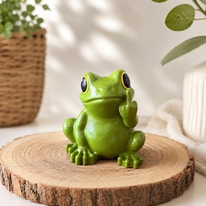 May include: A small green frog figurine with large black eyes and yellow rims. The frog is in a seated position with one hand raised, extending its middle finger upwards. The surface of the frog is smooth and painted in a vibrant shade of green.