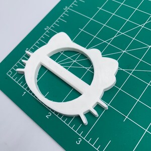 May include: White plastic belt buckle with a rounded, oval shape and a raised, horizontal bar in the center. The buckle has a textured surface and a slightly raised edge.