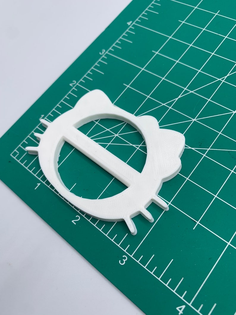 May include: White plastic belt buckle with a rounded, oval shape and a raised, horizontal bar in the center. The buckle has a textured surface and a slightly raised edge.