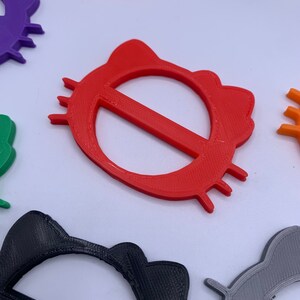 May include: A set of colorful plastic cat-shaped belt buckles. The buckles are in various colors, including red, purple, black, green, pink, gray, and orange. The buckles are all the same size and shape, with a cat's head and ears on the top and a circle in the middle.