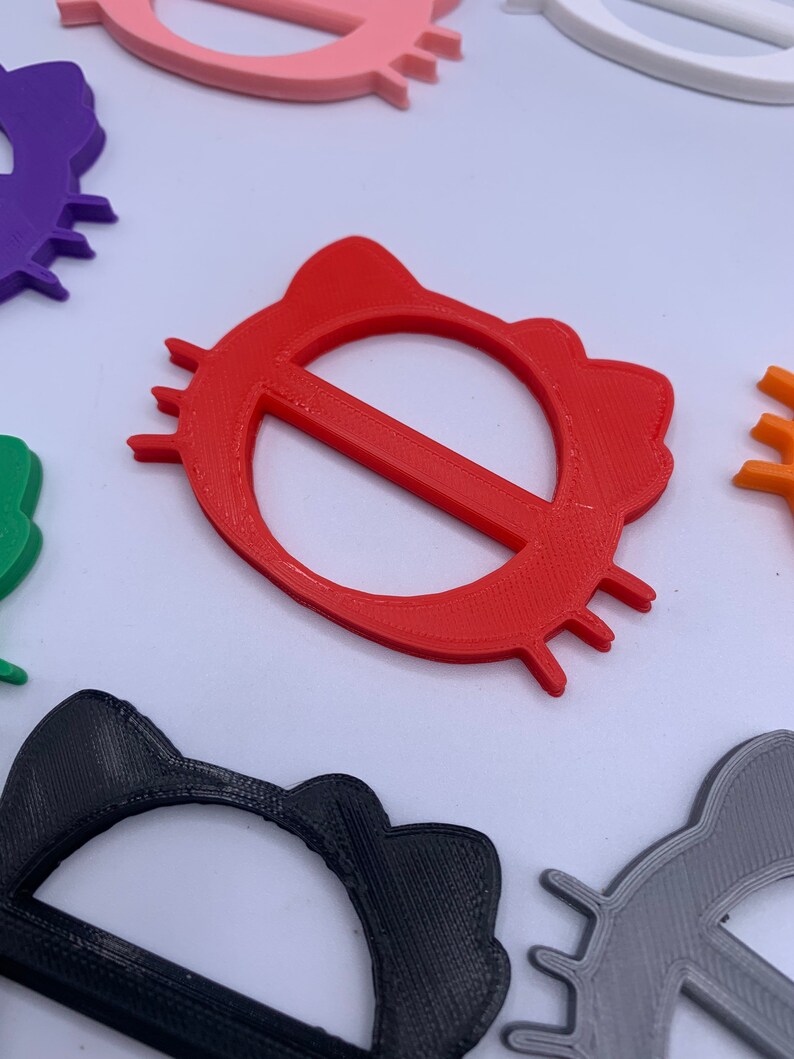 May include: A set of colorful plastic cat-shaped belt buckles. The buckles are in various colors, including red, purple, black, green, pink, gray, and orange. The buckles are all the same size and shape, with a cat's head and ears on the top and a circle in the middle.