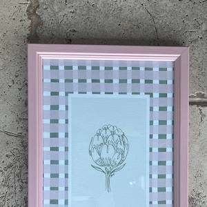 May include: A framed print of a green line drawing of an artichoke on a light blue background. The frame is pink with a white and green gingham pattern.