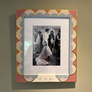 May include: A black and white photograph of a bride and her bridesmaids getting ready for a wedding. The bride is wearing a white wedding dress and a veil. The bridesmaids are wearing black dresses. The photo is framed in a pink and gold frame with a scalloped edge. The date "15.12.23" is written on the bottom of the frame.