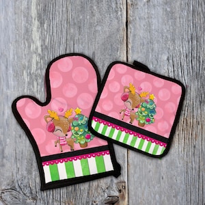 Puede incluir: Juego de manopla y agarradera rosas con temática navideña. El juego presenta un ciervo de dibujos animados con un árbol de Navidad sobre un fondo de lunares rosas. El borde inferior tiene rayas verdes y blancas.