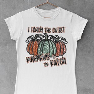 Peut inclure: T-shirt blanc avec le texte "J'enseigne les citrouilles les plus mignonnes du champ" et un graphique de trois citrouilles avec des accents de paillettes.
