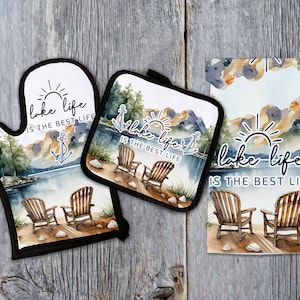 May include: A set of three kitchen items featuring a watercolor design of a lake scene with two chairs, a sun, and the text "lake life is the best life". The set includes a pot holder, an oven mitt, and a tea towel.