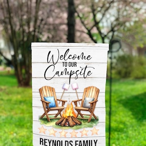 Könnte beinhalten: Ein weißes Holzschild mit dem Text "Welcome to our Campsite" in einer Schreibschrift. Das Schild zeigt zwei Holzstühle, ein Lagerfeuer und Marshmallows. Auf dem unteren Teil des Schildes steht "REYNOLDS FAMILY".
