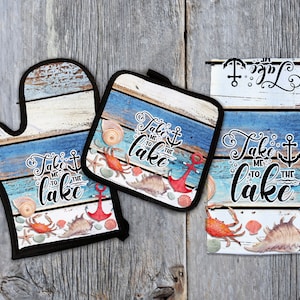 Puede incluir: Un conjunto de tres accesorios de cocina con un tema náutico azul y blanco sobre un fondo de tablones de madera. El conjunto incluye un posavasos, un guante para horno y una toalla de té. Cada artículo presenta el texto "Take me to the lake" con un ancla y conchas.